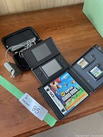 Nintendo DS Lite console open on a wooden surface, black carrying case with charger next to it, and a game case opened showing the New Super Mario Bros game box, two DS cartridges visible inside holder.