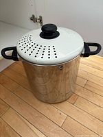 Large stainless steel pasta pot with white enamel perforated lid and black handles shown closed