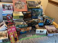 Multiple assorted jigsaw puzzle boxes stacked on a couch and floor showing a variety of images and brands.