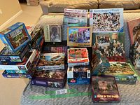 Photo showing a large assortment of boxed jigsaw puzzles stacked on carpet and bed, various brands and images visible, different puzzle counts from 750 to 2000 pieces.