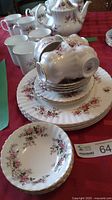 Set of lavender rose patterned fine china plates, cups and saucers with gold trim displayed on red tablecloth.