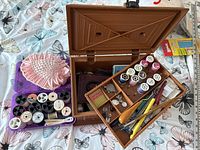 Wooden sewing box open showing threads, needles, measuring tape, scissors, and sewing tools inside compartments, a pink decorative pin cushion nearby.