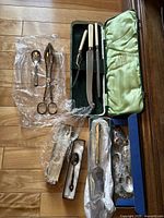 Photo of all items arranged on wooden floor including Cutlass carving set, silver serving tongs and spoon, and collector spoons wrapped in plastic and boxes.