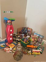 Photo showing a mix of toys, children's books, DVDs and video games including a red child's golf set, toy cars, and other collectibles on the floor against a beige wall.