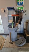Overview of the lot showing a gray plastic rolling garbage can with lid, a small sledgehammer, a shovel, a broom with orange bristles, and multiple black plastic and metal rakes and shovels leaning against a wall with an aluminum step ladder in the background.