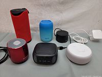 Photo showing six different Bluetooth speakers in red, blue, black, and white, some with cords and some without, positioned on a grey fabric surface.