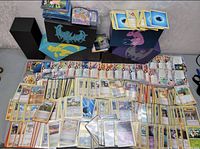 Wide view of a large number of Pokémon cards spread across a surface with several Pokémon-themed card boxes behind them, showing different card types and conditions.
