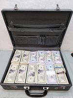 Open black briefcase displaying neatly arranged stacks of movie prop bills in multiple denominations appearing as fake US currency.