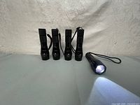 Five black compact LED flashlights with wrist straps arranged standing on a light surface. One flashlight is switched on showing bright white light beam.