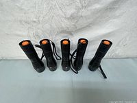 All five black LED flashlights with orange push buttons, lined up vertically suspended by wrist straps