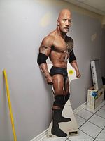 Full view of life-size standup cutout showing The Rock in wrestling gear with a muscular pose against a wall, beside household items for scale.