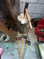 Three small axes on the floor in front of a log with one large wood splitter axe leaning against the log, placed in a basement or workshop floor setting.