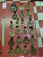 Full view of the 22 brass medallions arrayed on a red background, showing variety of shapes and attached leather straps.
