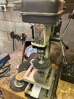 Front angle of the Brico drill press on workbench with abrasive polishing disc attached and various discs around.