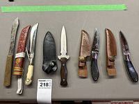 Overview of all knives arranged left to right: wood-handled carving knife, decorative knife with red sheath, double-edged dagger with black sheath, three Browning knives (two with leather sheaths, one loose)