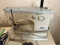 Front and side view of white and light blue Elna Super sewing machine with attached cords and pedal.