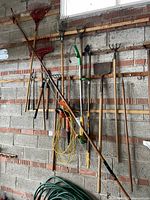 Photo showing assorted garden tools hanging on a wall including leaf rakes, pitchfork, shovels, hoes, an electric hedge trimmer, edger with power cord and other tools.
