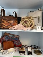 Photo showing medium brown leather handbag with geometric design, petit point floral bag, pink crocheted bag, beige leather handbag, and part of lower shelf with more jewelry and boxes.