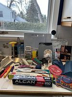 Wide view of assorted kitchen accessories on countertop including cutting boards, knives, graters, parchment and aluminum foil rolls, and scrubbing pads.