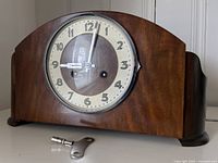 Front view of wooden mantle clock with circular cream dial and attached winding key on table in front.