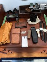 Top view of lot displaying men's tan leather gloves, three watches, leather wallets and pouch, mini binoculars on white cloth, a black digital alarm clock, cigar case, shoe brush, and vintage model car.