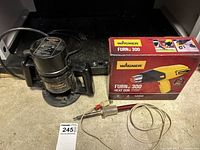 Photo of assembled Black & Decker plunge router, Wagner Furno 300 heat gun box, and Archer soldering tool placed on floor.