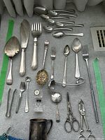 Assorted silver plate utensils spread out on a towel showing various spoons, forks, cake servers, scissors, tongs, ladle, and a metal cup.