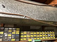 Long metal shelf with three black plastic multi-drawer caddies filled with assorted labeled small hardware drawers under ductwork in a basement environment