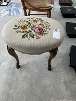 Close-up photo angled from above showing the embroidered needlepoint floral pattern on the beige stool upholstery and legs.