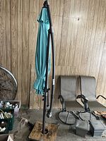 Full view of closed turquoise off-set patio umbrella with curved frame and square weighted base indoors against wood panel wall.