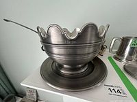 Full view of the pewter soup tureen with ladle resting inside and under plate underneath