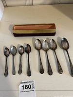 Nine silver plate spoons laid out in front of an open display box, varying in size and handle design, showing wear and tarnish.