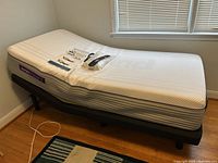 Twin adjustable bed with Ease brand black adjustable frame and Purple Premier 3 mattress in a small bedroom corner, mattress showing white quilted top with geometric pattern and purple side labels. Remote control, power cord, and manual on mattress.