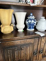 Four ceramic vases and urns on wooden shelf including a yellow vase with swans, white flared vase, blue and white urn with lid, and white lidded urn