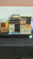 Lot arranged on black covered table including boxed UHF antennas, clock radios, telephones, tape-head cleaning kit, boxed indoor/outdoor speakers and other speakers