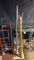 Photo showing red and blue metal dollies, branch trimmer with yellow handle, steel bar clamps, and metal rack and hanging shelf in background.