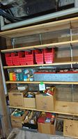 Wide view of shelving unit with red plastic bins on the top shelf and multiple cardboard boxes filled with hardware parts and tools below.