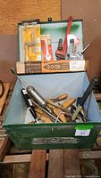 Green metal bin with assorted hand tools inside visible including hand saws, clamps, screwdriver, and caulking gun