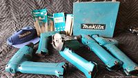 All five teal colored Makita cordless power tools visible with various attachments, accessories, Makita cases, manuals, and blue Makita baseball cap on black background.