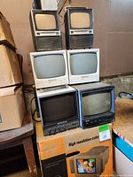 Seven vintage monitor units stacked and arranged on boxes including Panasonic, Yutek Shibaden, RCA and white CRT monitors.