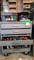 Full view of grey three-drawer metal workbench on wheels with plastic crate and case on top
