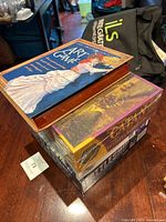 Four stacked board games including The Art Game, Catan Traders & Barbarians expansion, Kamisado, and Battleship on a wooden table with a fabric bag in the background.
