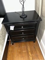 Front view of black wooden bedside table with three drawers and metal knobs.