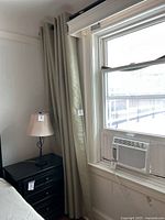 Light taupe blackout curtains hung beside a window with air conditioning unit. Curtains are pleated and floor-length, hanging from black curtain rod.