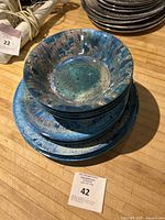Stack of melamine plates and bowls in blue/teal Radiance pattern with speckled finish arranged on a wooden surface.