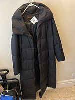Front view of the black Cole Haan puffer coat with padded collar on white hanger against wall, showing size tag.