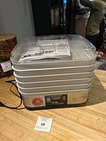 Front view of Hamilton Beach dehydrator showing digital control panel, stackable trays and clear lid with instruction manual inside.