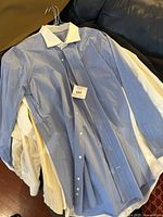Stack of men's dress shirts including a blue shirt on top with tag, placed on a couch.