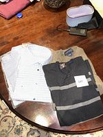 Photo showing three men's apparel items on a wood table: a tan sweater, a black and gray striped sweater and a white checkered dress shirt.