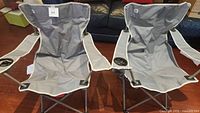 Two gray fabric folding chairs side by side with armrests and built-in cup holders